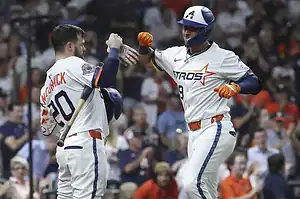 Two baseball players in Astros uniforms celebrate a successful play, one giving a fist bump while holding a bat. Crowd in the background.