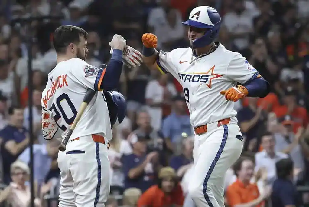 Two baseball players in Astros uniforms celebrate a successful play, one giving a fist bump while holding a bat. Crowd in the background.