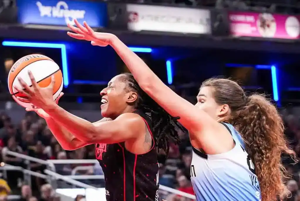 A basketball player in a red jersey attempts a layup while being closely defended by another player in a blue and white uniform.