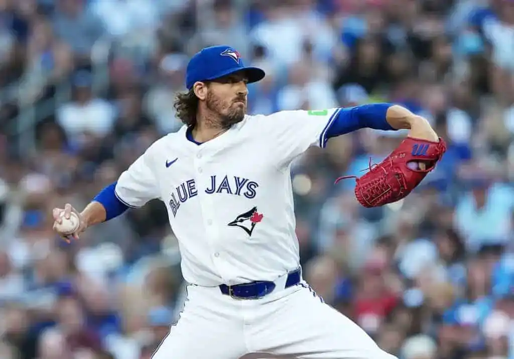 A pitcher in a white Toronto Blue Jays jersey throws a baseball, showcasing intense focus on the mound with a blurred crowd in the background.