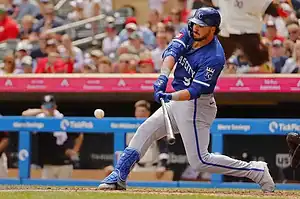 A baseball player in a blue uniform swings a bat to connect with a pitch, with a crowd of fans visible in the background.