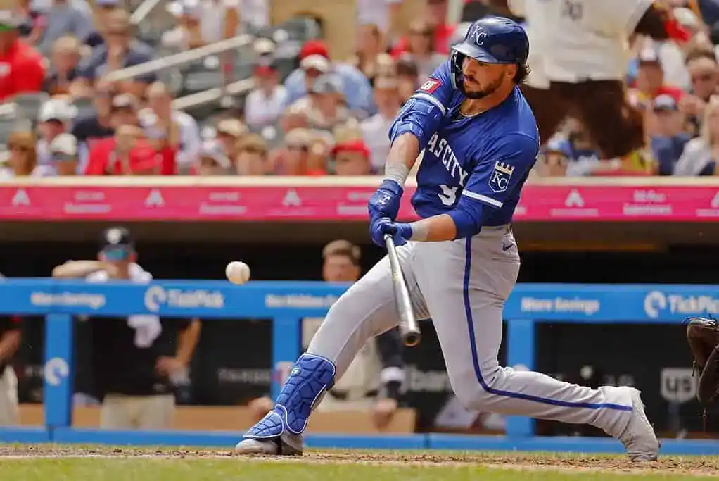 A baseball player in a blue uniform swings a bat to connect with a pitch, with a crowd of fans visible in the background.