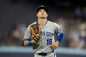 A baseball player in a gray jersey with the text "Kansas City" is running on the field, wearing a glove on his left hand.