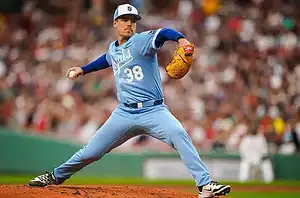 A baseball pitcher in a light blue uniform throws a pitch on a vibrant field, surrounded by a cheering crowd.
