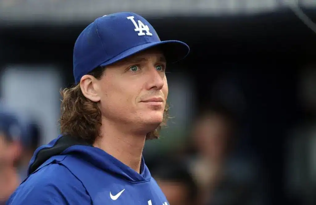 A baseball player in a blue Dodgers cap and hoodie stands in a dugout, focused amidst a blurred background of teammates.