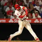 A baseball player in a red and white uniform swings at a pitch, with excited fans watching in the background at a game.