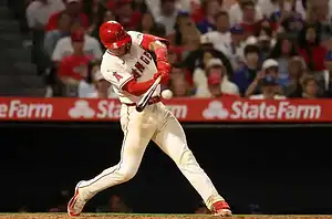 A baseball player in a red and white uniform swings at a pitch, with excited fans watching in the background at a game.