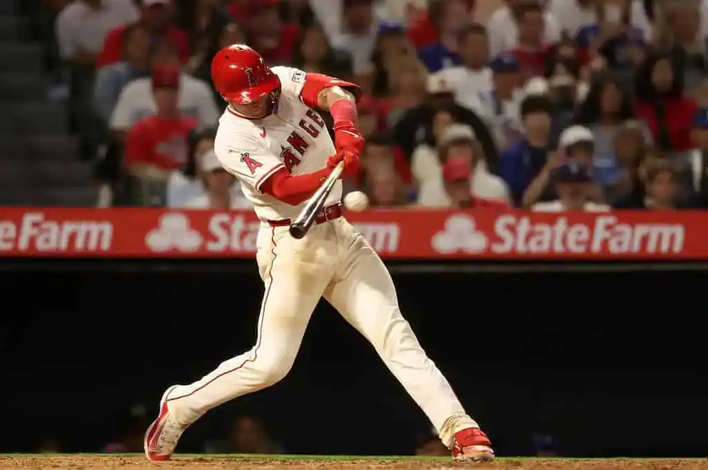 A baseball player in a red and white uniform swings at a pitch, with excited fans watching in the background at a game.