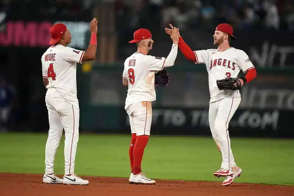 Three baseball players in white and red uniforms from the Angels celebrate together on the field, sharing high fives and camaraderie.