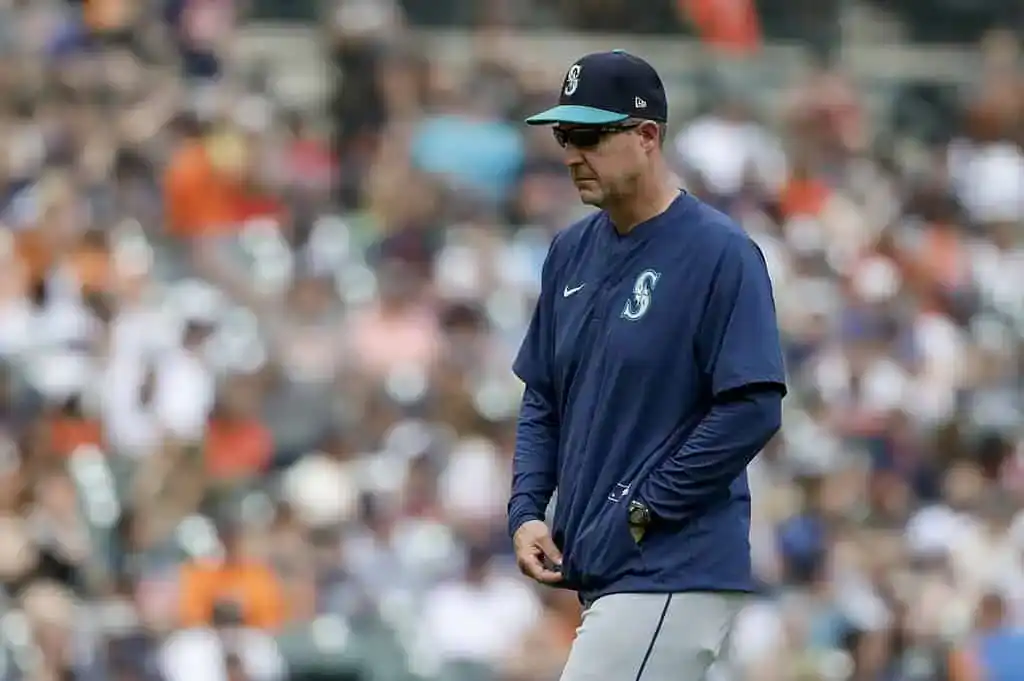 A Seattle Mariners coach wearing a navy cap and sunglasses walks on the field during a game.