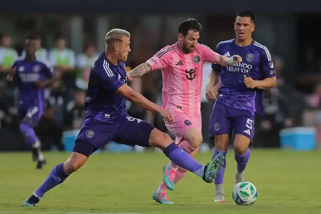 Two players in purple uniforms attempt to tackle a player in a pink jersey during an intense soccer match on the field.