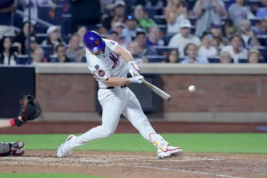A baseball player in a white and blue uniform swings at a pitch, with crowds visible in the stands behind him at a night game.