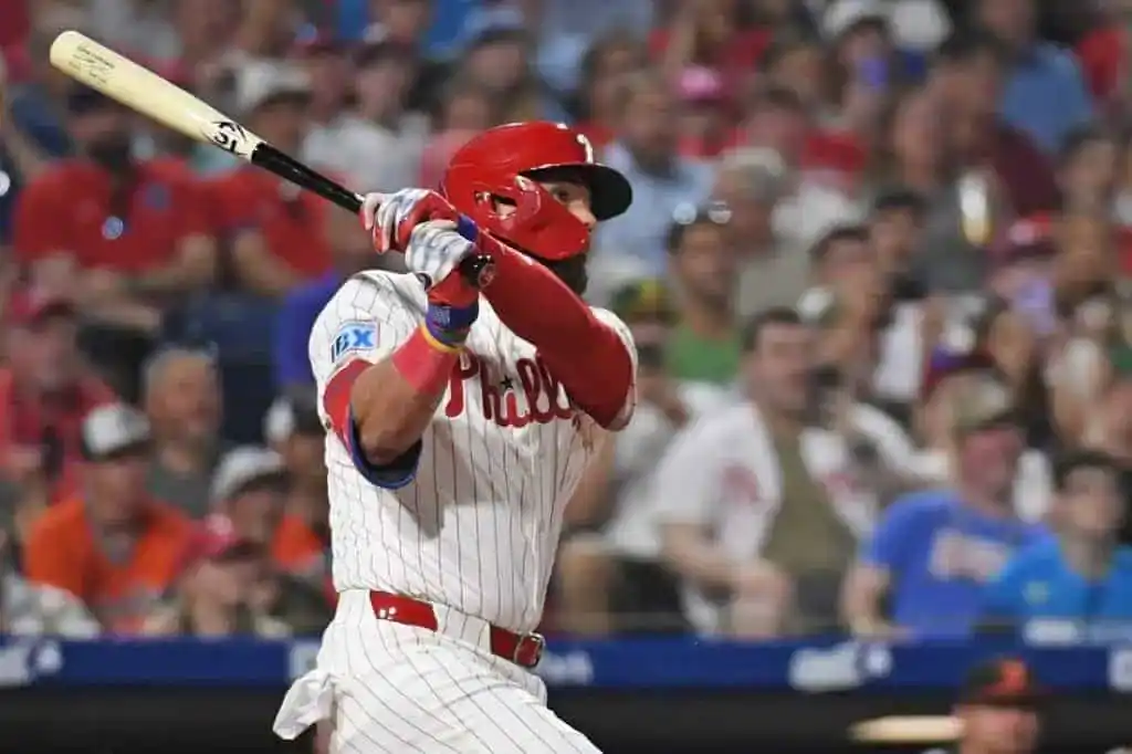 A Philadelphia Phillies player swings a baseball bat, with fans cheering in the background at a lively baseball game.