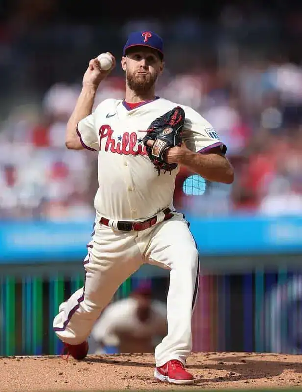A Philadelphia Phillies pitcher winds up to throw a baseball during a game, wearing a white jersey and red cleats on the mound.