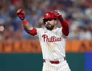 A Philadelphia Phillies player celebrates on the field, wearing a white striped jersey with red accents and raising his arms excitedly.