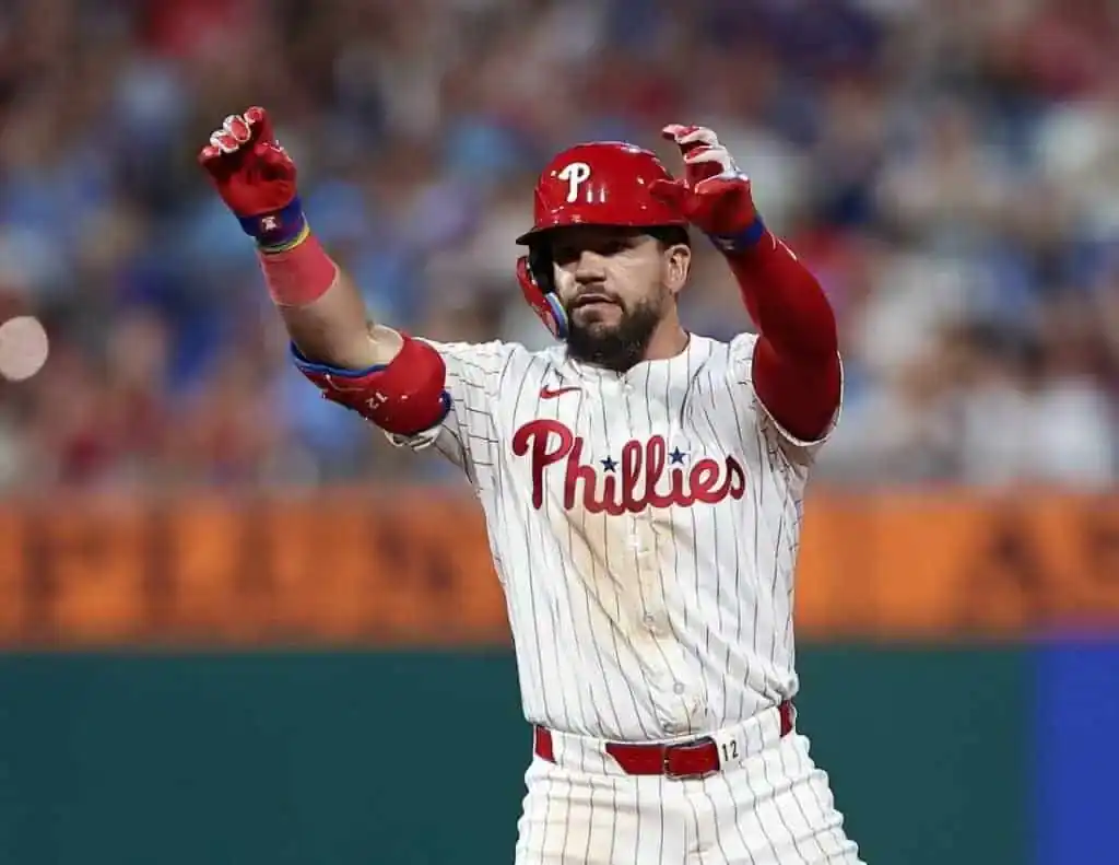 A Philadelphia Phillies player celebrates on the field, wearing a white striped jersey with red accents and raising his arms excitedly.