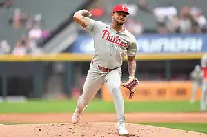 A pitcher in a gray Phillies uniform winds up to throw a pitch on a baseball field, with spectators visible in the background.