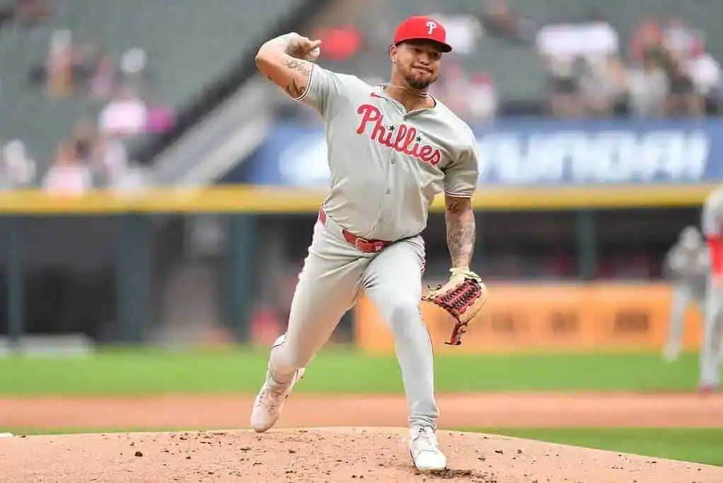 A pitcher in a gray Phillies uniform winds up to throw a pitch on a baseball field, with spectators visible in the background.