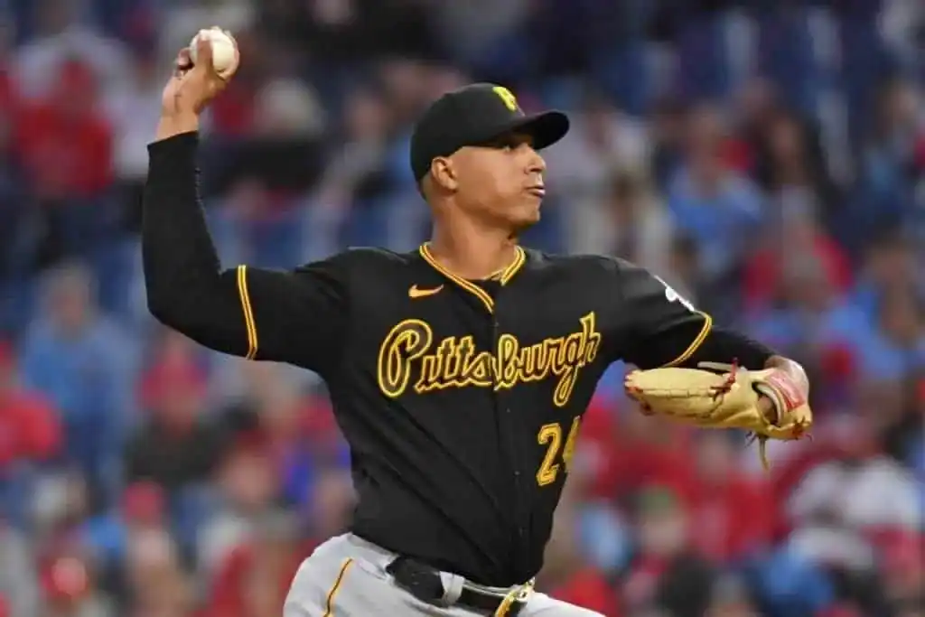 A Pittsburgh Pirates player in a black jersey prepares to throw a baseball, with the stadium and fans blurred in the background.