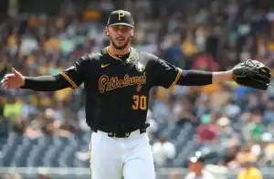 A pitcher in a black and gold Pittsburgh Pirates jersey, arms outstretched, stands on the mound in a busy stadium.