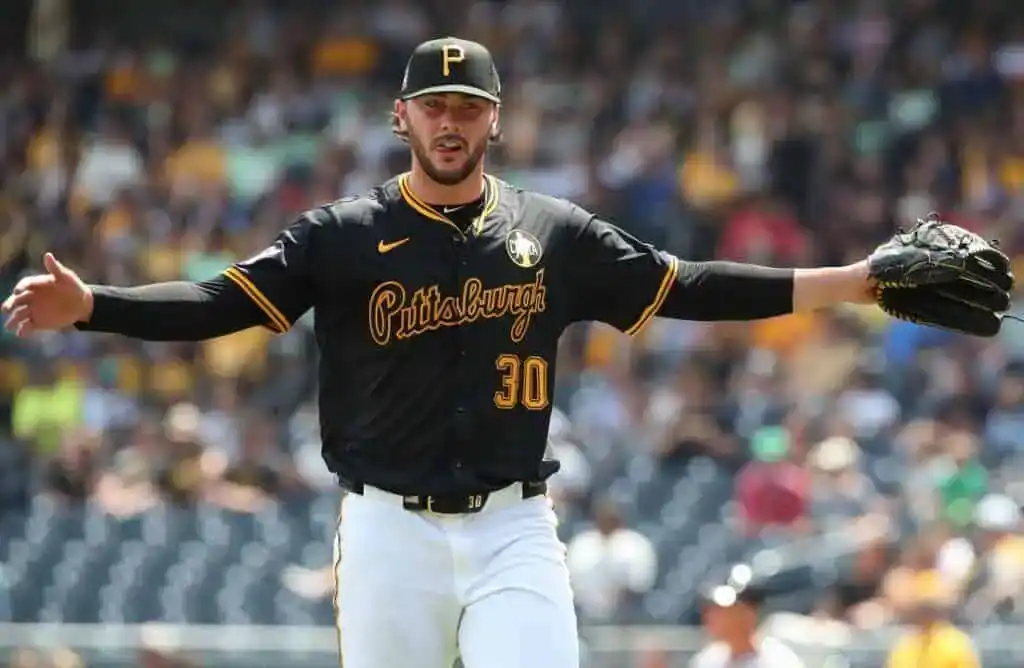 A pitcher in a black and gold Pittsburgh Pirates jersey, arms outstretched, stands on the mound in a busy stadium.
