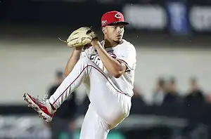 A baseball pitcher in a white uniform with red accents prepares to throw a pitch on a dimly lit field.