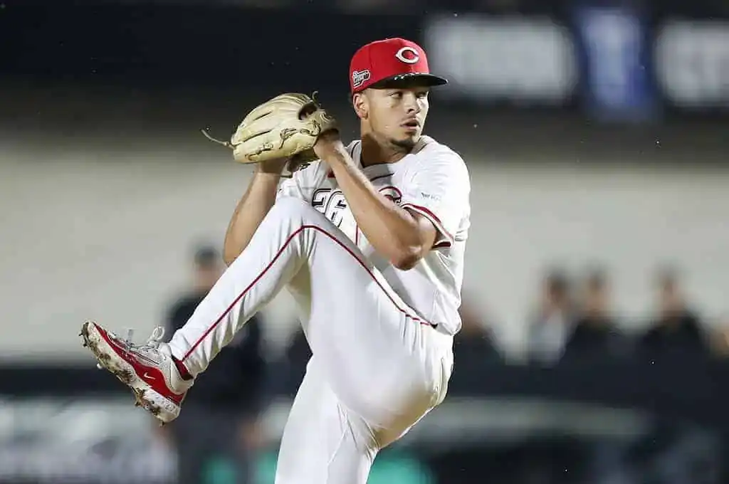 A baseball pitcher in a white uniform with red accents prepares to throw a pitch on a dimly lit field.