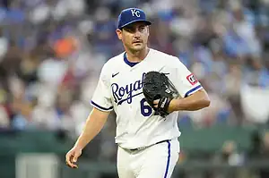A Kansas City Royals player in a white uniform and navy cap holds a baseball glove, walking on the field during a game.