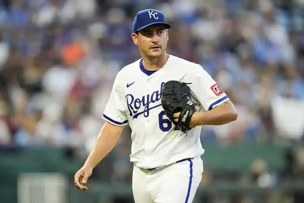 A Kansas City Royals player in a white uniform and navy cap holds a baseball glove, walking on the field during a game.