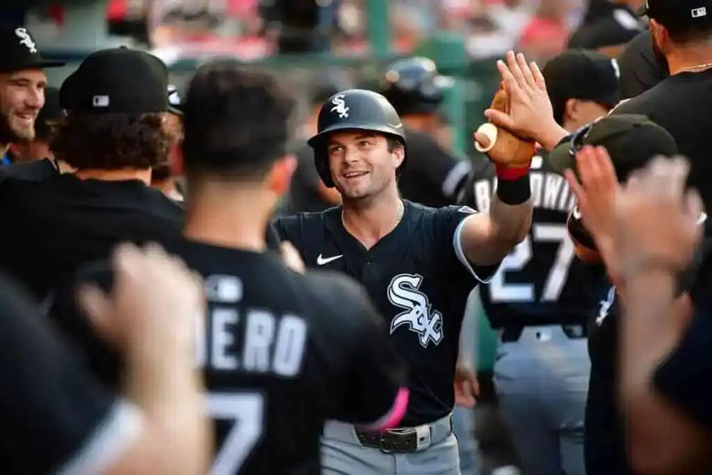 A baseball player in a black jersey celebrates with teammates in a dugout, high-fiving and holding a bat, showcasing team camaraderie.