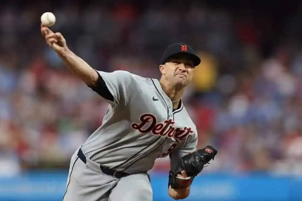 A baseball pitcher in a gray Detroit Tigers uniform winds up to throw a baseball during a game, with spectators blurred in the background.
