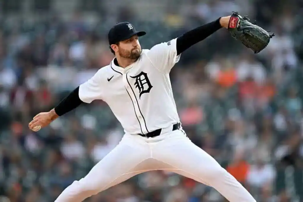 A baseball pitcher in a Detroit Tigers uniform, poised in a throwing stance on the field, with a blurred stadium crowd in the background.