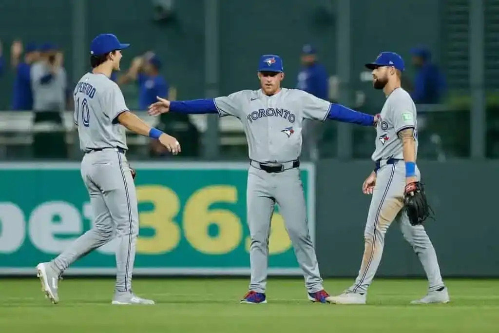 Three Toronto Blue Jays players in grey uniforms engage in a lively discussion on the field during a baseball game.