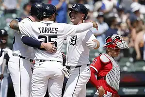 Baseball players in white jerseys celebrate a hit with hugs, while a catcher in red watches from behind the plate during a game.