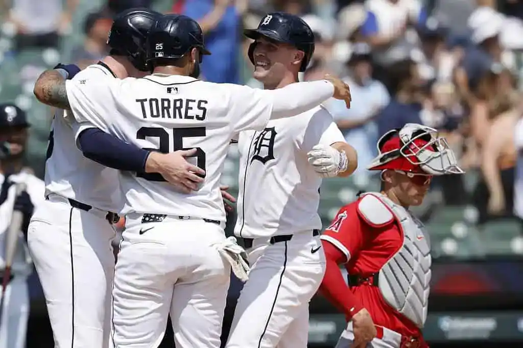 Baseball players in white jerseys celebrate a hit with hugs, while a catcher in red watches from behind the plate during a game.