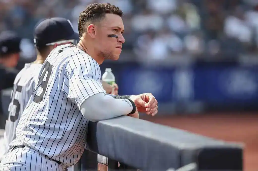 A baseball player in a pinstriped uniform leans on the dugout railing, observing the game in a sunlit stadium.