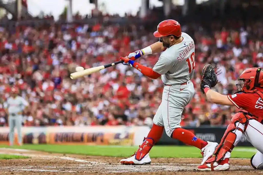 A baseball player swings at a pitch, with a catcher poised to receive the ball, amidst a crowd filled with enthusiastic fans.