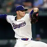 A Texas Rangers pitcher in a white uniform winds up to throw a baseball during a game, showcasing action and intensity on the field.