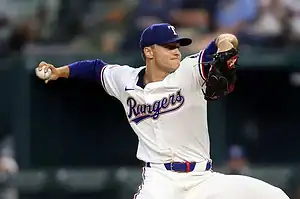 A Texas Rangers pitcher in a white uniform winds up to throw a baseball during a game, showcasing action and intensity on the field.