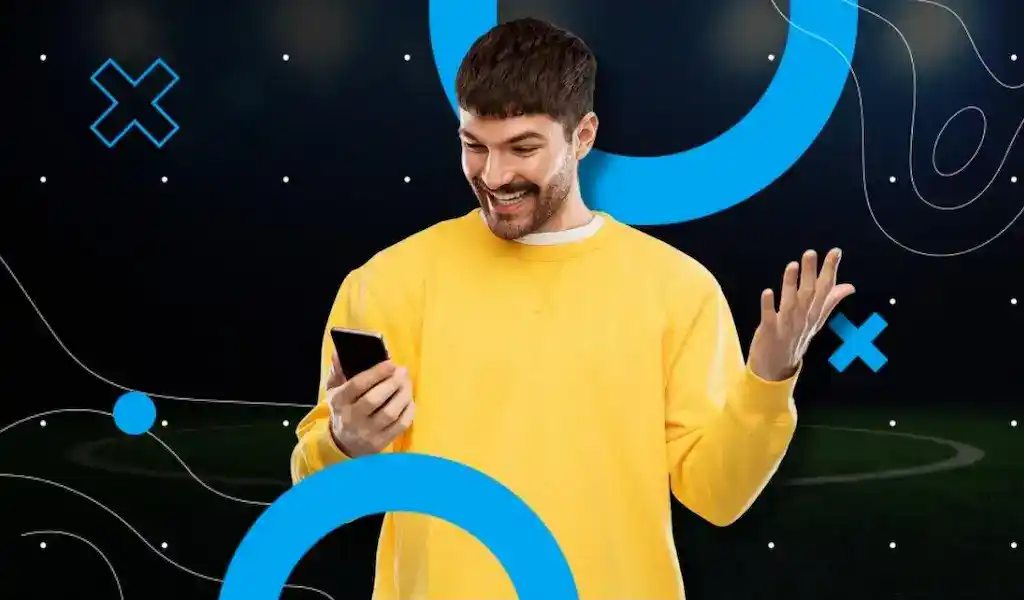 Smiling man in yellow sweatshirt checking his phone on a soccer field background, symbolizing how odds guide fans and analysts in sports betting.