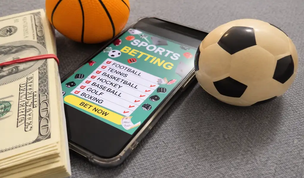 Smartphone showing a sports betting app beside cash, a basketball, and a soccer ball, illustrating technology and user experience in mobile sports betting.